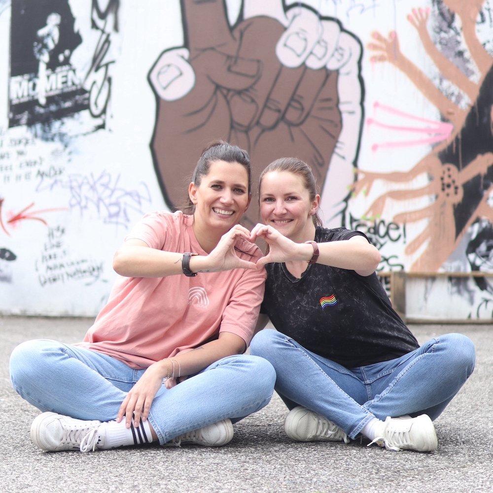 Jasmin und Steffi sitzen nebeneinander auf dem Boden vor einem Graffiti und formen ein Herz mit den Händen