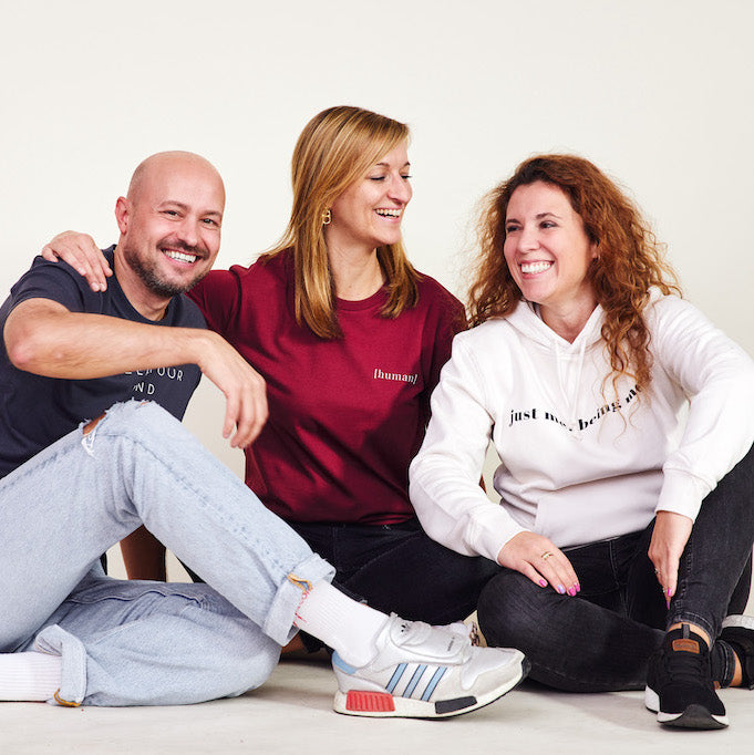 Drei Personen sitzen auf dem Boden, lächeln zueinander und tragen Kleidung von BeeProud