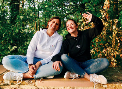 Die beiden Gründerinnen sitzen auf einer Mauer vor einem Wald. Beide schauen lachend in die Kamera. Steffi, die rechte Person winkt zusätzlich.