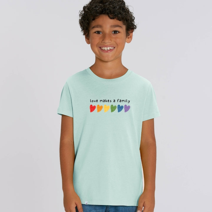 Kind lächelt in die Kamera und trägt karibikblaues Shirt mit dem Aufdruck love makes a family und bunten Herzen