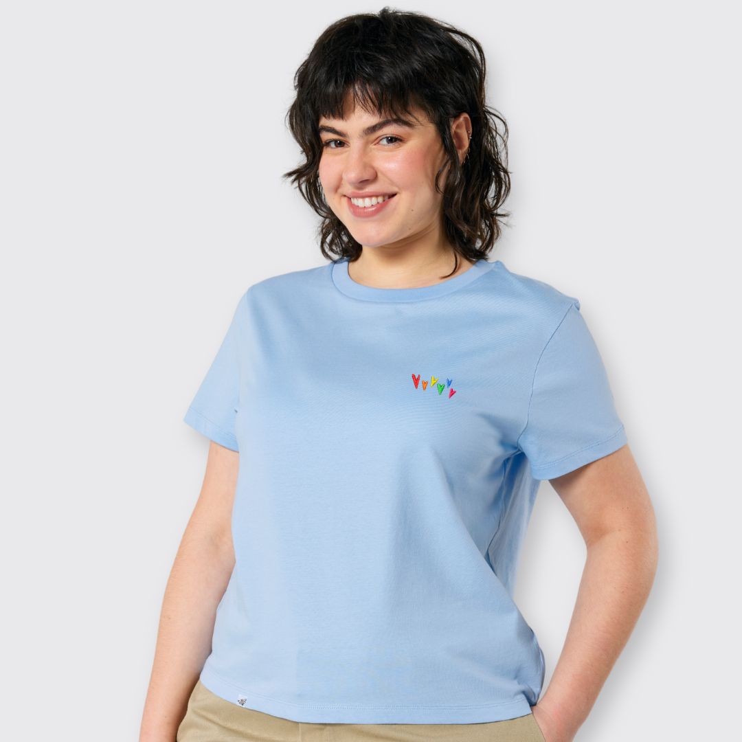 Model lächelt und trägt hellblaues Shirt mit kleinen gestickten Herzen in Regenbogenfarben