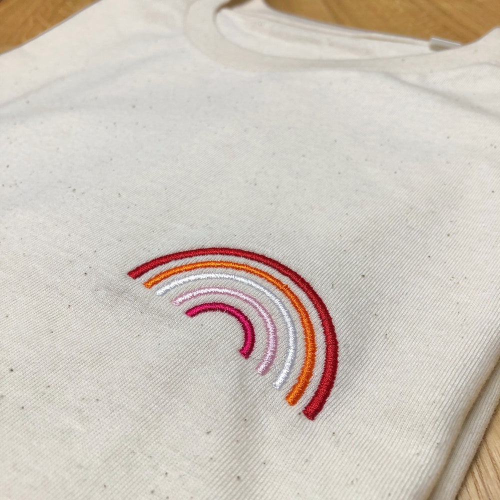 Nahaufnahme naturfarbenes Shirt mit Regenbogen Stick in Farben der lesbischen Flagge