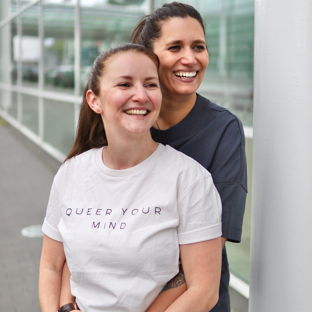 Zwei Personen die lachen, eine davon trägt ein cremefarbenes Shirt mit der Aufschrift queer your mind