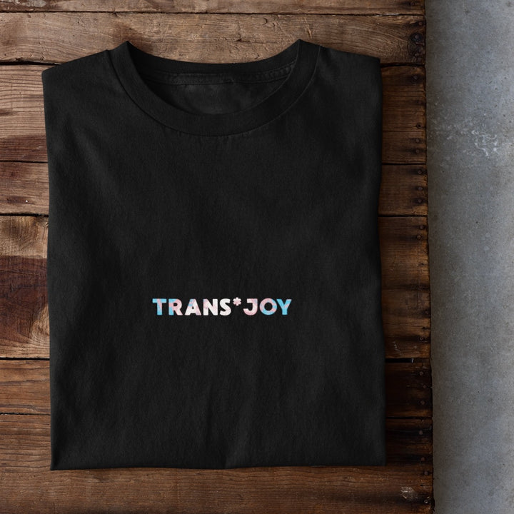 Schwarzes Shirt mit dem mittigen Aufdruck transjoy in den Farben hellblau, rosa und weiß liegt zusammengefaltet auf einem Holztisch