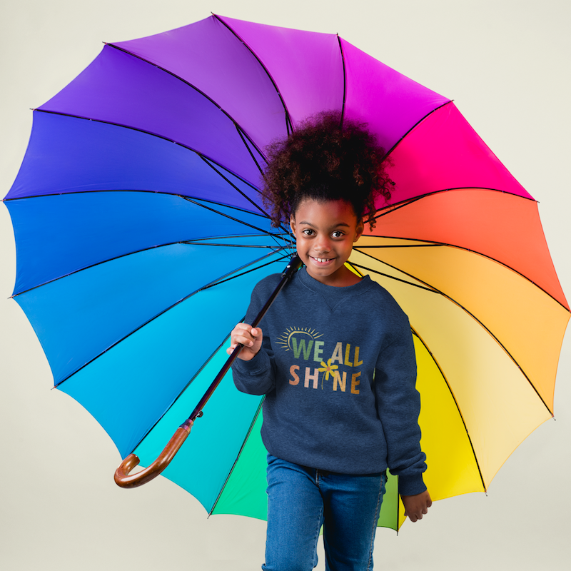 Kind hält einen regenbogenfarbenen Regenschirm und trägt einen dunkelblauen Pullover mit dem Aufdruck we all shine