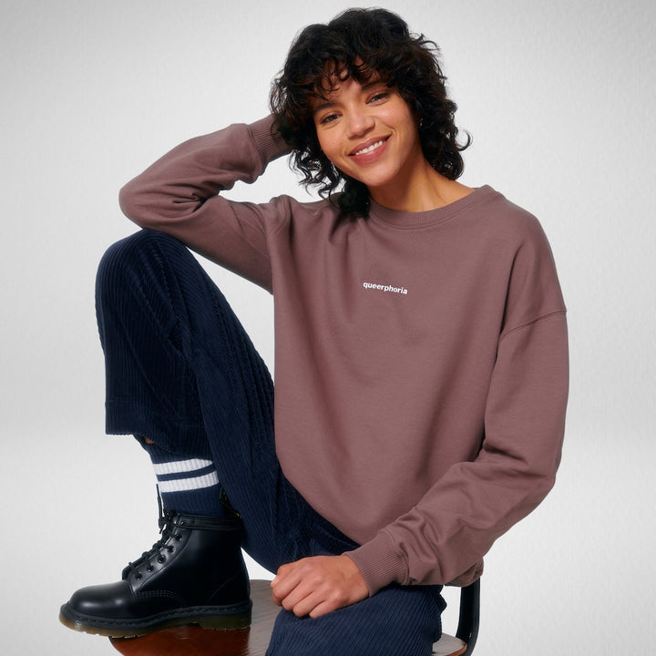 Person sitzt auf einem Hocker, lächelt in die Kamera und trägt ein Oversized Sweatshirt in der Farbe Aubergine mit dem Stick queerphoria