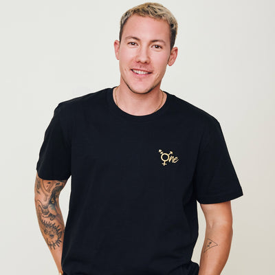 Brix Schaumburg trägt schwarzes Shirt mit dem All Gender Symbol ONE in goldenem Stick